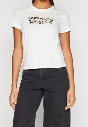 Femme portant un t-shirt blanc à manches courtes avec un logo imprimé léopard beige et noir, associé à un jean noir taille haute.