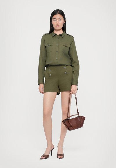 Olivnozeleni srajcni zgornji del s epauleti na ramenih, usklajeni kratki hlače z dvojnimi gumbi, v kombinaciji z rjavim usnjeno torbico in sandali z odprtimi prsti.