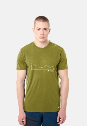 Giovane uomo con capelli corti che indossa una maglietta Vaude verde oliva con design di montagna bianca e pantaloni blu, in piedi davanti a uno sfondo chiaro.