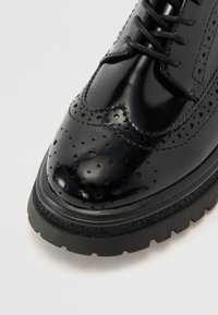 Black patent leather shoe with brogue detailing, featuring a smooth, glossy surface and a thick rubber sole. Laces are included.