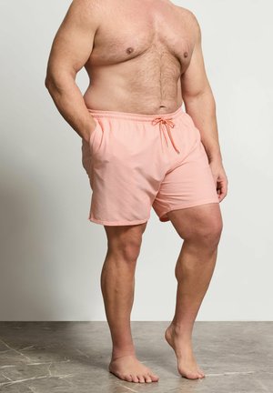 Hombre sin camiseta con bañador rosa claro a rayas, con las manos en los bolsillos, de pie descalzo sobre un suelo de mármol gris.