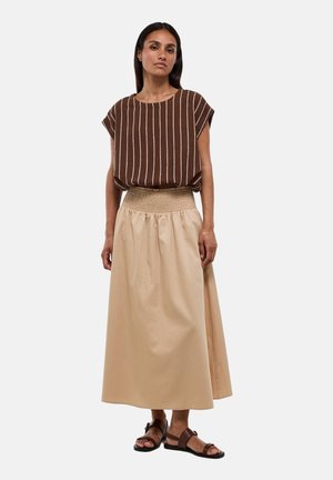 Femme debout portant un haut marron rayé à manches courtes, une longue jupe beige taille haute et des sandales plates marron foncé sur un fond uni.