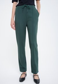 Zielone, rozciągliwe spodnie z wiązaniem w pasie, kieszeniami bocznymi, pionowymi szwami oraz dopasowanym krojem, w połączeniu z czarnymi, płaskimi butami.