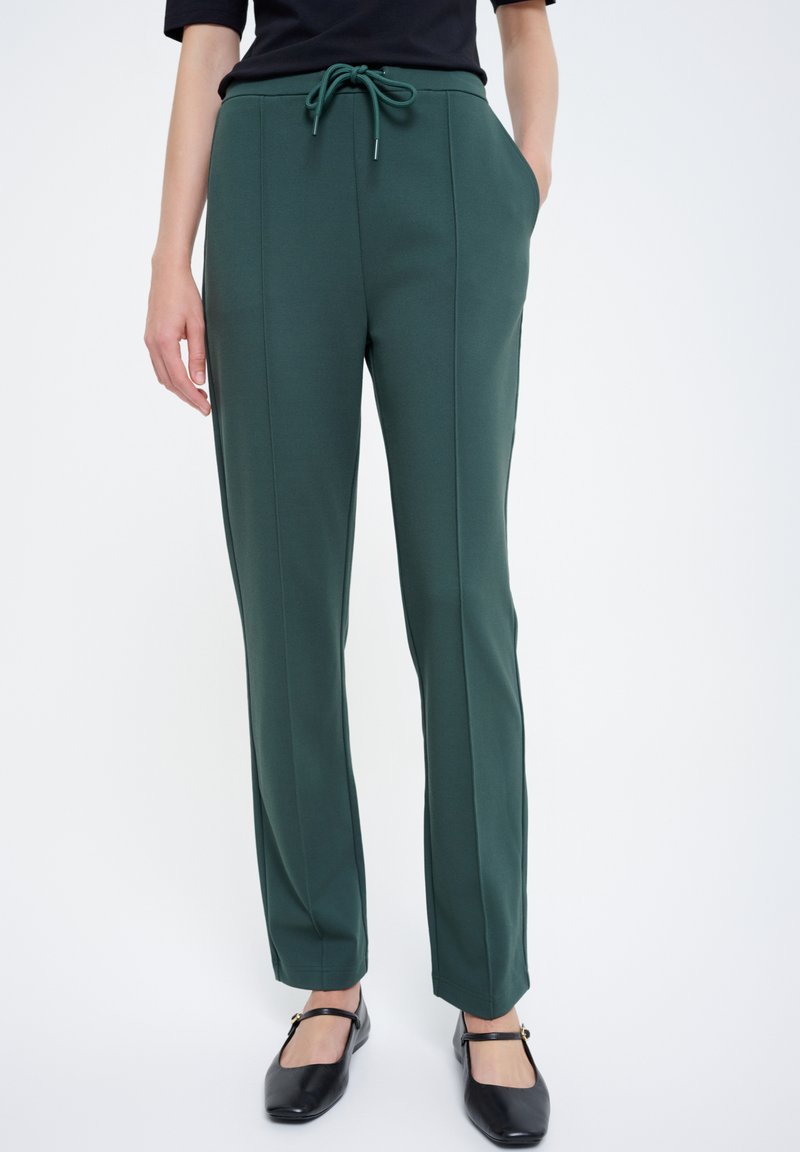 Zielone, rozciągliwe spodnie z wiązaniem w pasie, kieszeniami bocznymi, pionowymi szwami oraz dopasowanym krojem, w połączeniu z czarnymi, płaskimi butami.