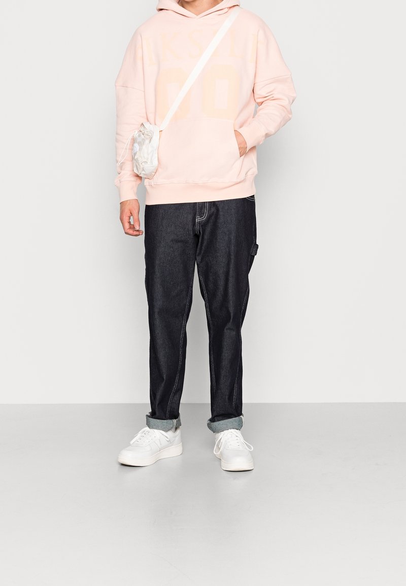 Jasnoróżowa bluza z kapturem z dużym nadrukiem, ciemne dżinsy z podwiniętymi mankietami, białe trampki oraz mała biała torebka noszona przez ramię.