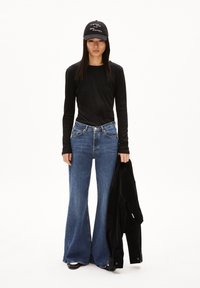 Schwarzes Langarmoberteil, blaue weite Jeans, schwarze Mütze mit Text, hält eine schwarze Jacke, steht vor einem schlichten weißen Hintergrund.