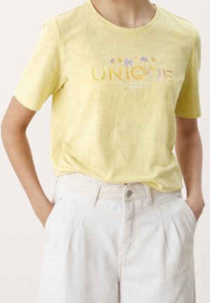 Osoba nosząca żółtą koszulkę z napisem "UNIQUE" i kwiatami nadrukowanymi ponad białymi spodniami z wysokim stanem, ręce w kieszeniach.