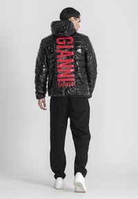 Uomo girato di spalle che indossa un giubbotto a vento nero lucido con testo rosso "GIANNI KAVANAGH" sulla schiena, pantaloni neri e sneakers bianche.