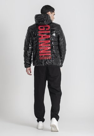 Chaqueta acolchada negra y brillante con capucha, que presenta un destacado texto del logo en rojo en la parte posterior. Combinada con pantalones negros y zapatillas blancas.