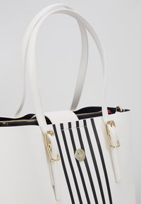 Sac à main blanc avec des rayures verticales noires à l'avant, ferrures dorées, double poignée et une fermeture éclair visible à l'intérieur.