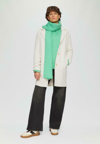 Manteau crème avec un col, pull en laine verte, écharpe teal, pantalon noir surdimensionné et baskets beiges avec une semelle marron.