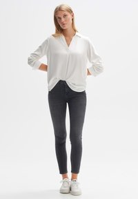 Blusa bianca a maniche lunghe con scollatura a V, vestibilità rilassata e polsini arrotolati, abbinata a jeans skinny scuri e sneakers bianche. Design minimalista.
