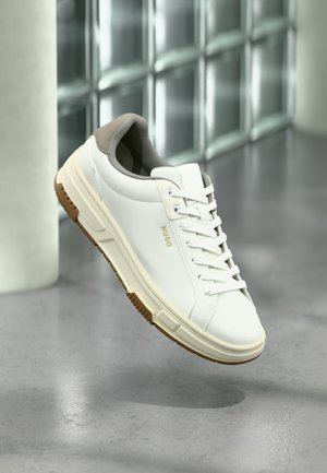 Baskets blanches avec des accents beige, semelle en caoutchouc et lacets, de marque "HUGO", posées au-dessus d'un sol en béton poli près de blocs de verre.