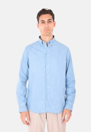 Uomo che indossa una camicia a maniche lunghe azzurro chiaro abbottonata e pantaloni beige, in piedi su uno sfondo grigio chiaro uniforme.