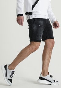 Pantalones cortos de mezclilla negra con un aspecto desgastado, puños enrollados y manchas de pintura blanca. Combinados con zapatillas deportivas en blanco y negro.