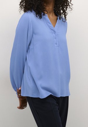 Femme aux cheveux bouclés portant un chemisier bleu clair texturé à manches longues avec des boutons et un pantalon foncé, debout, les mains dans le dos.
