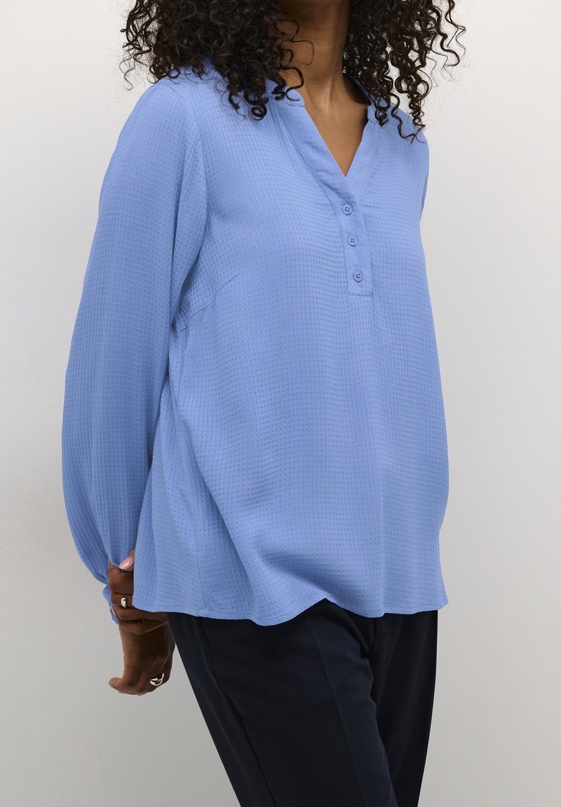 Femme aux cheveux bouclés portant un chemisier bleu clair texturé à manches longues avec des boutons et un pantalon foncé, debout, les mains dans le dos.
