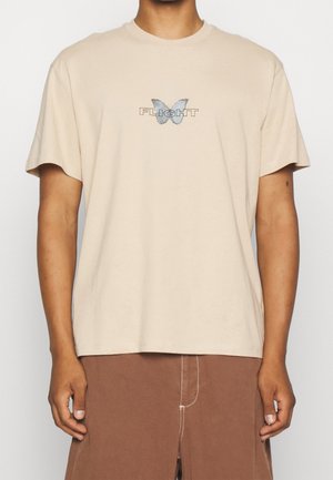 Beżowy t-shirt z krótkim rękawem z niebieską grafiką motyla i napisem "FLIGHT" na piersi, noszony przez osobę w brązowych spodniach.