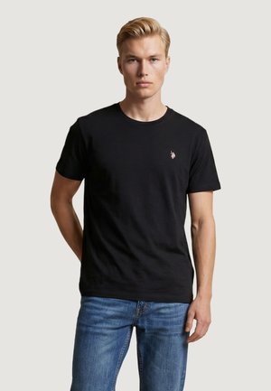 Giovane uomo con capelli biondi corti che indossa una t-shirt nera a maniche corte con un piccolo logo sul petto e jeans blu, in piedi davanti a uno sfondo semplice.