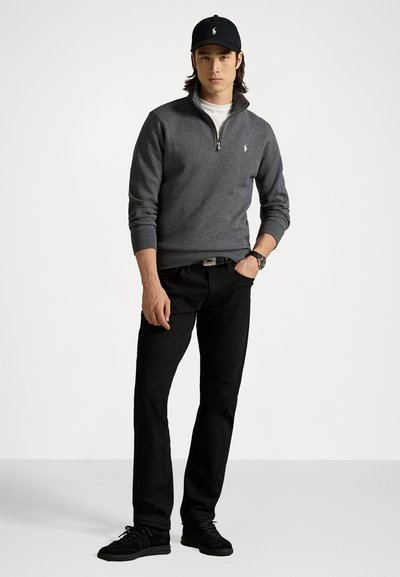 Grijze zip-up trui van zachte stof, gecombineerd met zwarte jeans en zwarte sneakers. Geaccessoireerd met een zwarte pet en horloge.