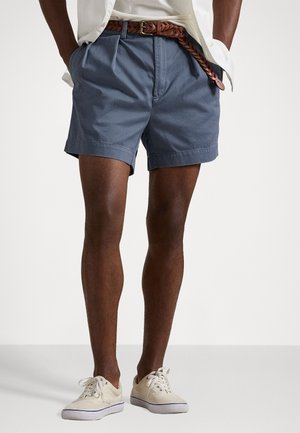 Shorts gris pour homme avec des plis, une ceinture tressée marron et des baskets en toile blanches. Les shorts sont en tissu léger et ont une coupe ajustée.
