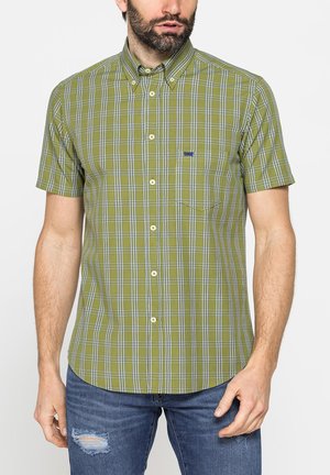 Uomo che indossa una camicia verde oliva a maniche corte con motivo a quadri bianco e blu, abbinata a jeans blu strappati.