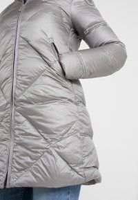 Een persoon in een lichtgrijze gewatteerde jas met ritszakken en hand in een zak tegen een effen achtergrond.