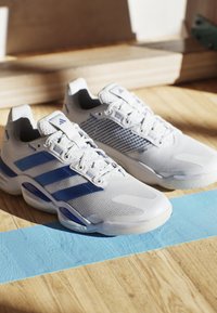 Białe buty sportowe z niebieskimi akcentami, siateczkowym wzorem i wyściełanym kołnierzem. Posiadają trzy niebieskie paski po bokach oraz teksturowaną podeszwę.