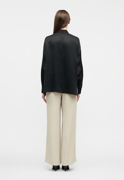 Γυναίκα με μακριά καστανά μαλλιά, βλέποντας από πίσω, φοράει μια φαρδιά μαύρη μπλούζα με δαντελένια μανίκια και μπεζ παντελόνες με φαρδιά γραμμή, συνδυασμένες με μαύρα τακούνια.