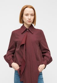 Blouse rayée bordeaux avec un col pointu, un devant plissé et des manches longues. Le tissu a une texture lisse et une coupe décontractée.