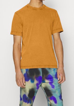 Camiseta mostaza de manga corta con escote redondo, que presenta una textura suave y lisa, y un dobladillo sin acabar. Combinada con leggings coloridos y estampados.