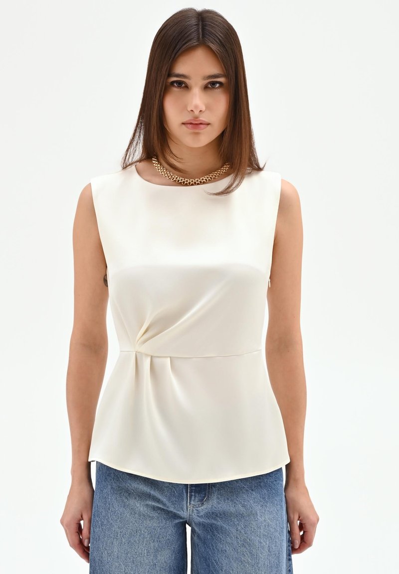 Jonge vrouw met stijlbruin haar draagt een mouwloze witte blouse met plooi-detail en blauwe jeans, geaccentueerd met een gouden ketting.