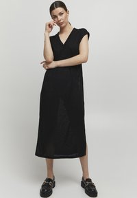Abito nero, lungo fino al ginocchio, con scollatura a V e maniche corte, realizzato in un materiale a maglia testurizzato e con spacchi laterali. Abbinato a mocassini neri.
