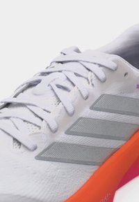 Λευκό αθλητικό παπούτσι με γκρι ρίγες, λευκά κορδόνια και φωτεινή πορτοκαλί σόλα, εμφανιζόμενο σε κοντινή λήψη εστιάζοντας στην περιοχή των κορδονιών.