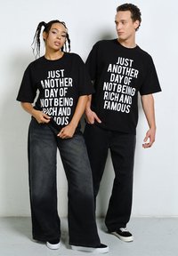 Two young adults pose wearing black t-shirts with white text that reads, "Just another day of not being rich and famous."