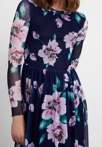Robe florale avec une base bleu marine ornée de grandes fleurs roses et de feuilles vertes, manches longues en voile transparent, et jupe froncée.
