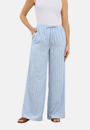 Vrouw draagt een wijde broek met verticale blauw-witte strepen en een taille met trekkoord, gecombineerd met een witte mouwloze top. Ze staat met één hand in haar zak.