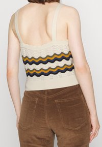 Béžový pletený top s vlnitými okraji a vícebarevnými zvlněnými pruhy (námořnická modř, olivová, zlatá) nad hnědým manžetem z kordurové látky.
