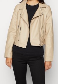 Beige bikerjack van imitatieleer met een cropped ontwerp, ritssluitingen op de zakken en zilverkleurige metalen details. Draagt over een zwarte top.