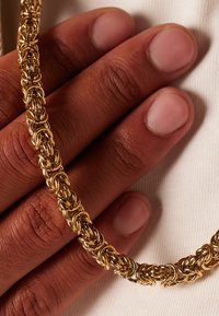 Gouden ketting met ingewikkeld gevlochten schakels, rustend op een hand met lichte huid. De gladde textuur en glans benadrukken het vakmanschap.