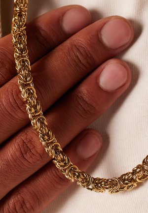 Gouden ketting met ingewikkeld gevlochten schakels, rustend op een hand met lichte huid. De gladde textuur en glans benadrukken het vakmanschap.