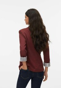 Blazer bordeaux à manches trois-quarts, avec des poignets blancs rayés. Tissu lisse avec une silhouette ajustée, porté avec un jean foncé.
