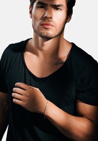 Svart T-skjorte med korte ermer båret av en mann, som viser en sølv armbånd på håndleddet hans. Han har lys hud og et tatovering på kravebeinet.