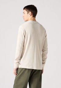 Beige langermet skjorte med ribbestruktur, rund halslinning og tettsittende mansjetter, sammen med olivengrønne bukser. Liten logo på sømmen.