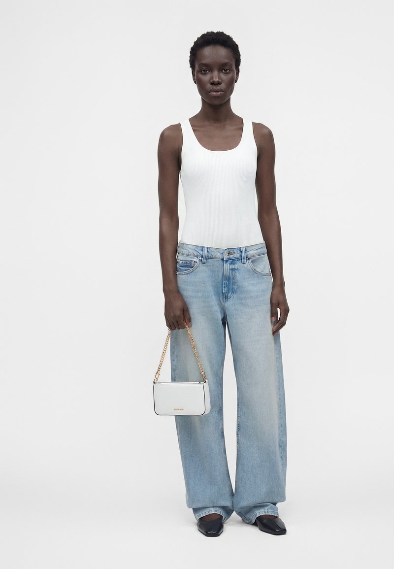 Femme portant un haut blanc sans manches, un jean large bleu clair, des chaussures noires, tenant un petit sac blanc avec une chaîne dorée.
