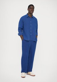 Man stands with hands in pockets wearing a blue button-up shirt, matching loose pants, and white sneakers against a plain white background.