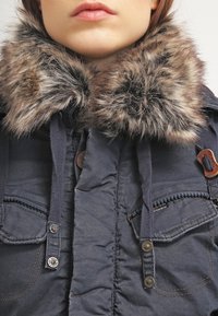 Mörkblå jacka med en mjuk, konstpälskrage, dragkedjefickor och knappar. Har ett texturerat tyg med sömnadsdetaljer.