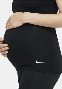 Czarny dopasowany top i legginsy z białym logo Nike, ukazujące ciążowy brzuch trzymany dłonią. Gładka tekstura i elastyczny materiał.