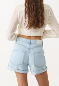 Vaaleansiniset farkkushortsit, joissa on käännetyt lahkeet, kaksi takataskua ja merkki vyötäröllä. Yhdistetty läpikuultavaan valkoiseen puseroon.