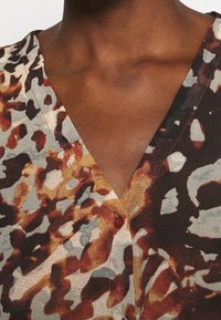 T-shirt med kort ärm och v-ringning i ett flerfärgat abstrakt mönster i brunt och grått, med bruna knappar vid halslinningen och en mjuk textur.